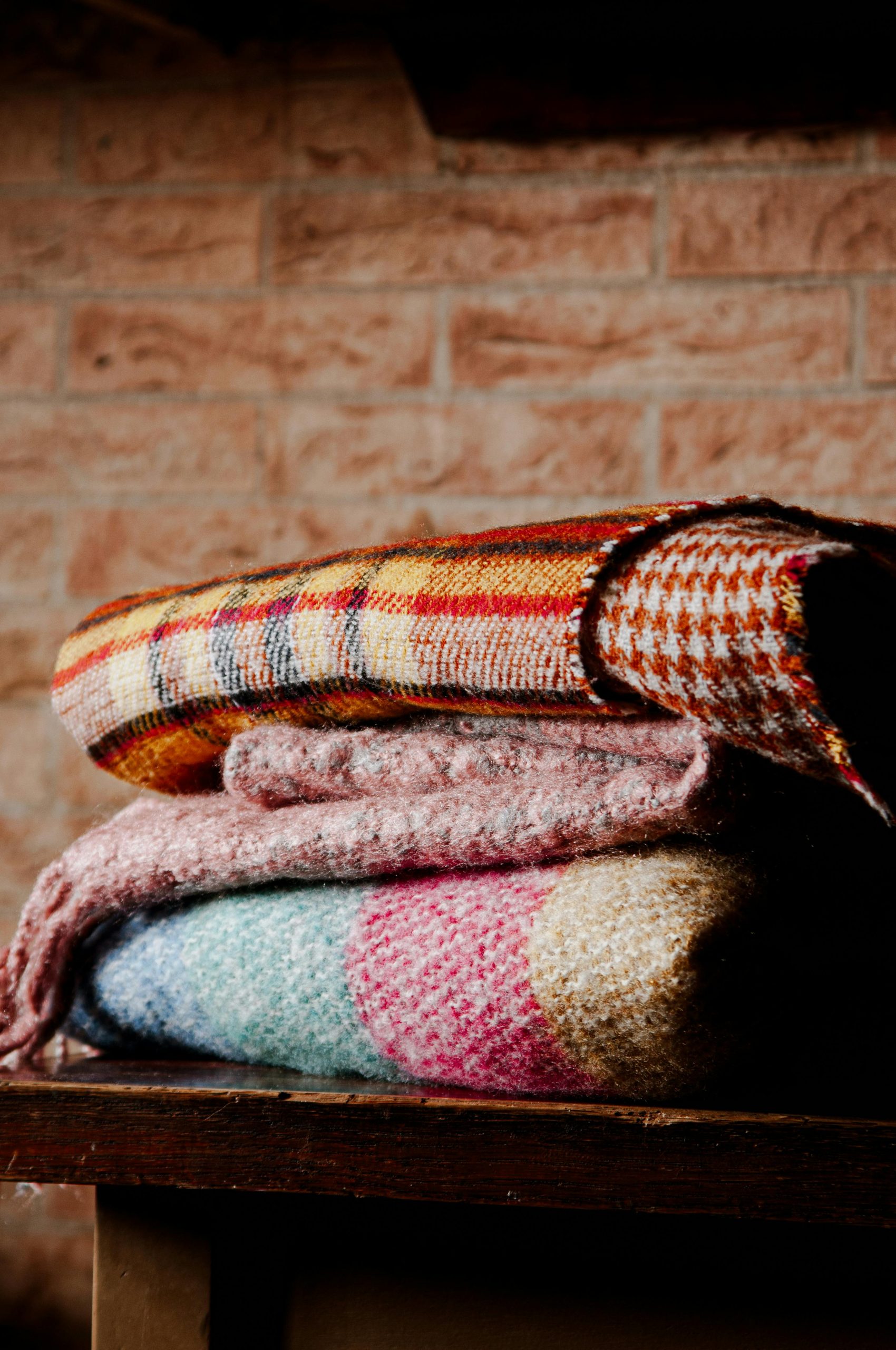 Stacked colorful blankets on a wooden table against a brick wall indoors.